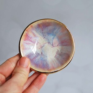 K&ouml;nnte beinhalten: Eine kleine, runde Keramikschale mit einer wirbelnden, mehrfarbigen Glasur in Rosa-, Blau- und Cremet&ouml;nen. Die Schale hat einen unregelm&auml;&szlig;igen, organischen Rand und ist mit einem metallisch goldenen Rand versehen, der den gesamten Umfang umschlie&szlig;t.