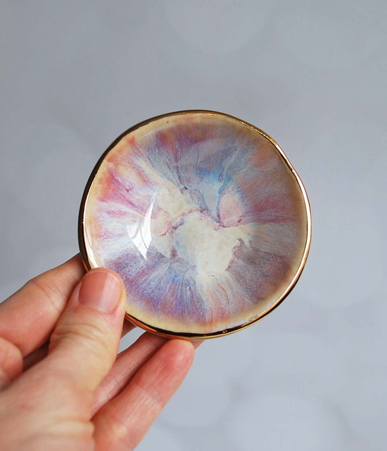 K&ouml;nnte beinhalten: Eine kleine, runde Keramikschale mit einer wirbelnden, mehrfarbigen Glasur in Rosa-, Blau- und Cremet&ouml;nen. Die Schale hat einen unregelm&auml;&szlig;igen, organischen Rand und ist mit einem metallisch goldenen Rand versehen, der den gesamten Umfang umschlie&szlig;t.