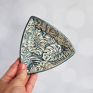 May include: A triangular ceramic dish with a teal glaze and a white and gold leaf pattern. The dish is perfect for holding jewelry, keys, or other small items.
