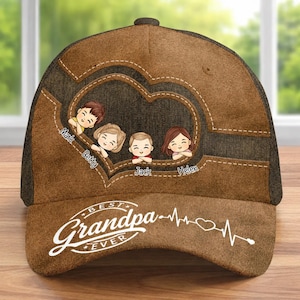 Pode incluir: Boné de beisebol castanho com um design em forma de coração com figuras de desenhos animados e nomes. O boné tem o texto "Best Grandpa Ever" em branco, juntamente com um gráfico de batimento cardíaco. O boné é feito de tecido castanho com uma parte traseira em malha.