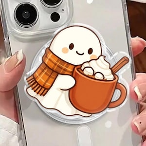 Può includere: Custodia trasparente per telefono con un fantasma dei cartoni animati con una sciarpa arancione e marrone, che tiene in mano una tazza marrone di panna montata e un cucchiaio. La custodia è su un telefono argentato.