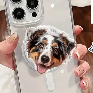 May include: Clear phone case with a custom dog face pop socket. The pop socket features a colorful image of a dog's face with the text "CUSTOM FACE". The phone case is held by a person.