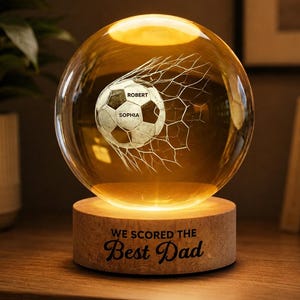 May include: A glowing glass sphere with a soccer ball design inside, set on a wooden base. The soccer ball is in a net, with the names "ROBERT" and "SOPHIA" on it. The base reads "WE SCORED THE Best Dad". The sphere is lit from within.