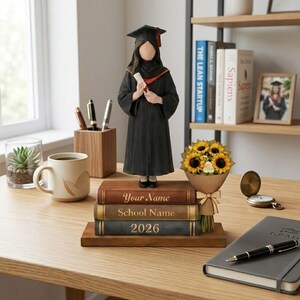 May include: A graduation figurine of a person in a black cap and gown, holding a diploma. The figurine stands on a stack of books with personalized text. A bouquet of sunflowers and a coffee mug are also on the desk.