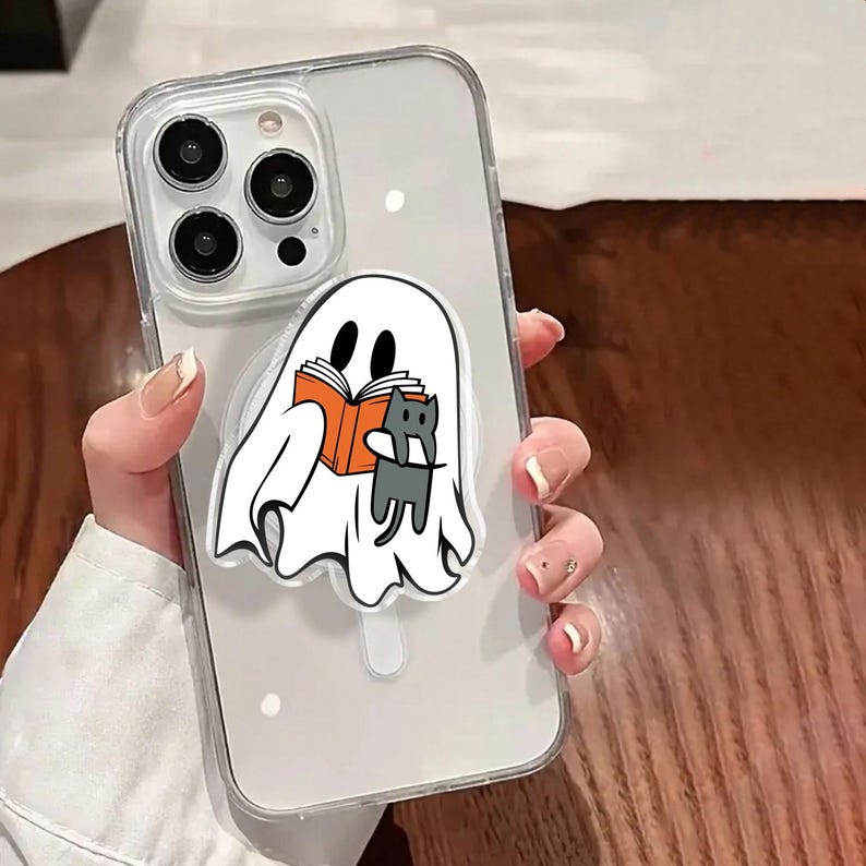 May include: Clear phone case with a white ghost design reading a book with a gray cat. The ghost and cat are holding an orange book. The phone case is clear with a white border.