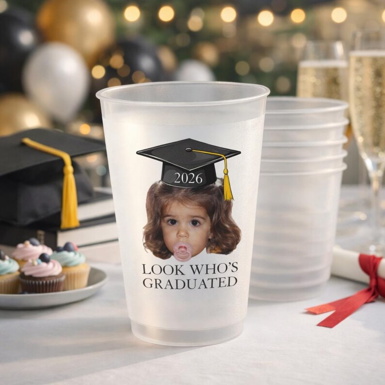 May include: Frosted plastic graduation party cups. Each cup features a graphic of a child wearing a graduation cap with the year 2026 and the text "LOOK WHO'S GRADUATED". Other cups, cupcakes, and champagne flutes are in the background.