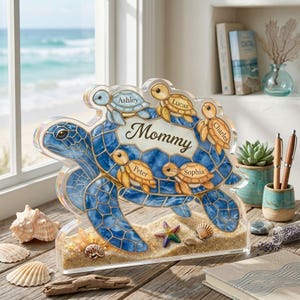 Op de afbeelding: Decoratief blauw zeeschildpadbeeldje met het woord "Mommy" en de namen Ashley, Lucas, Charles, Peter en Sophia. Het beeldje heeft een met zand gevulde basis met schelpen en een zeester. De achtergrond toont een raam met uitzicht op de oceaan.