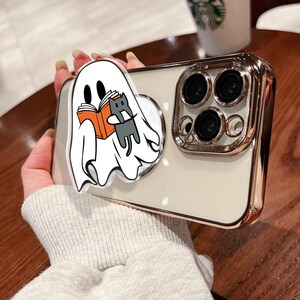 May include: A smartphone case with a gold-colored frame and a white back. A pop-up grip features a cartoon ghost reading a book with a gray cat. The case has a gold-colored frame and a white back.