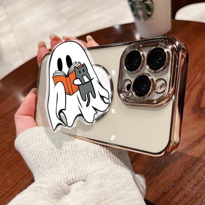 May include: A smartphone case with a gold-colored frame and a white back. A pop-up grip features a cartoon ghost reading a book with a gray cat. The case has a gold-colored frame and a white back.