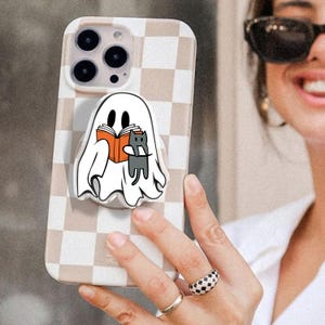May include: A phone case with a beige and white checkered pattern. A pop-up grip features a cartoon ghost reading a book with a gray cat. The phone case is held by a person wearing sunglasses and rings.