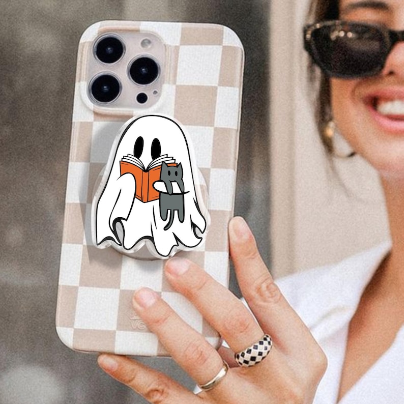 May include: A phone case with a beige and white checkered pattern. A pop-up grip features a cartoon ghost reading a book with a gray cat. The phone case is held by a person wearing sunglasses and rings.