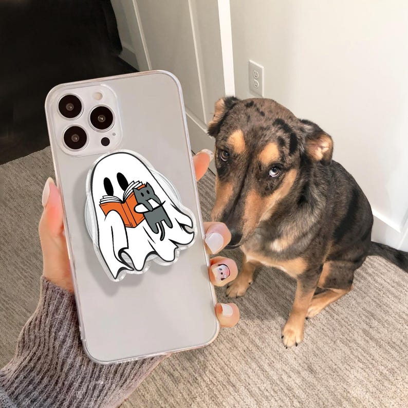 May include: A clear phone case with a white ghost-shaped phone grip. The grip features a cartoon ghost reading a book with a gray cat. A dog with brown and black fur is in the background.