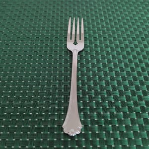 May include: A silver fork with four tines, a decorative handle, and a heart-shaped cutout. The fork is set against a green woven placemat.