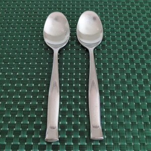 May include: Two stainless steel spoons with oval bowls and elongated handles. The spoons are positioned side by side against a green woven background. The spoons have a polished, reflective surface.