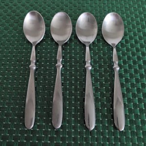 May include: Four stainless steel spoons with oval bowls and decorative handles are arranged in a row. The spoons are set against a green woven placemat. The spoons are a classic design, suitable for everyday use.