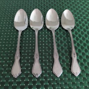 May include: Four silver-toned metal spoons arranged in a row. Each spoon features a polished oval bowl and a handle decorated with a delicate embossed floral pattern and a beaded border along the edges, ending in a rounded, flared base.