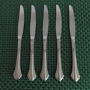 Puede incluir: Cinco cuchillos de mesa de acero inoxidable sobre una superficie verde texturizada. Los cuchillos tienen una hoja curva y un mango decorativo con un diseño acanalado.