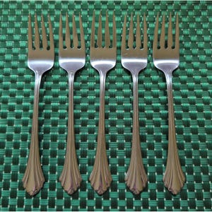 May include: A set of five identical stainless steel dinner forks arranged in a row. Each fork features a polished metallic finish, four tines, and a decorative handle base with vertical fluted grooves that flare out toward the bottom edge.