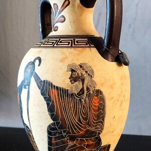 Puede incluir: Un jarrón de cerámica color crema con asas y base marrón oscuro. El jarrón presenta una ilustración en blanco y negro de una figura sosteniendo un pájaro. Patrones y detalles decorativos adornan el cuello.