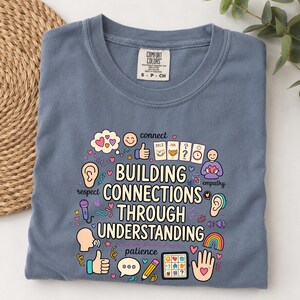 Könnte beinhalten: Ein schieferblaues T-Shirt mit dem Text "BUILDING CONNECTIONS THROUGH UNDERSTANDING", umgeben von bunten Illustrationen wie Sprechblasen, Herzen, einem Regenbogen, einem Bleistift, Händen, Ohren und verschiedenen Symbolen zur Kommunikation.