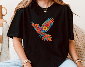 Puede incluir: Camiseta negra con un vibrante diseño floral de pájaro. El pájaro está compuesto por flores coloridas en tonos rojos, naranjas, amarillos, azules y morados. La camiseta tiene mangas cortas y cuello redondo.