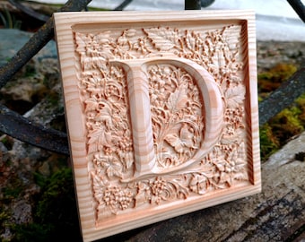 May include: A wooden plaque featuring a carved letter "D" surrounded by intricate floral designs. The plaque is made of light wood and has a natural finish.
