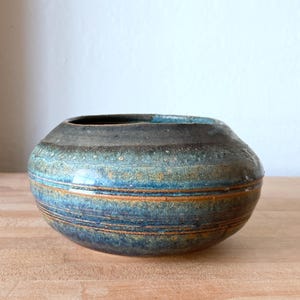 May include: A round ceramic bowl with a wide opening, featuring a dark charcoal rim that transitions into a vibrant blue and earthy brown glaze. The surface displays horizontal bands and a textured, mottled finish across the rounded body.