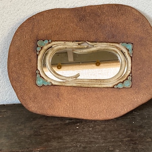 May include: A handcrafted, organic-shaped mirror with a textured, brown ceramic frame. The mirror has a cream-colored, oval-shaped inner frame with turquoise accents. Two small, gold-colored knobs are visible on the mirror's surface. The mirror is placed on a wooden surface.
