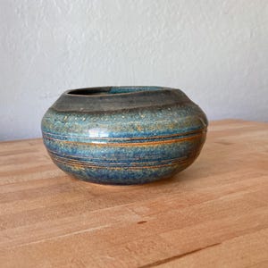 May include: A handcrafted ceramic vase with a round shape and a wide opening. The vase features a textured surface with horizontal bands of blue, teal, and orange. The vase is approximately 5 inches in diameter and 4 inches tall.