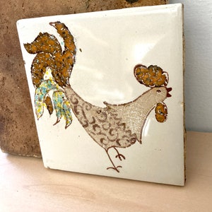 May include: A square ceramic tile with a hand-painted rooster design. The rooster features brown, tan, and blue accents against a cream-colored background. The tile is approximately 6 inches square.