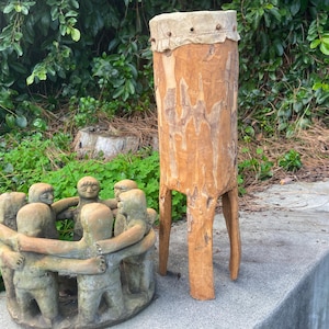 May include: A handcrafted wooden drum with a natural finish and a stretched hide top. Beside it, a sculpture of figures holding hands in a circle. The drum stands on three legs, and the sculpture is a light brown color.