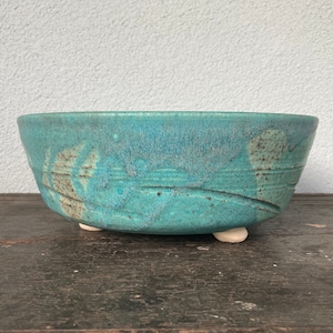 May include: A handcrafted, turquoise ceramic bowl with a textured surface and three small, white feet. The bowl features a wide, shallow design with subtle, organic patterns and is suitable for decorative or functional use.