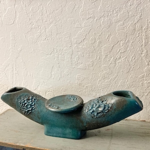 May include: A teal ceramic vase with a unique, asymmetrical design. The vase features two openings on either end and a circular accent in the center. The surface has textured, raised floral patterns and a dark, aged appearance. The vase is displayed on a weathered wooden surface.