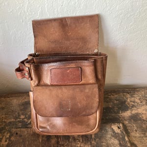 May include: A brown leather tool pouch featuring a large main compartment with a fold-over flap, a smaller front pocket with a flap closure, and a side loop. The leather shows a weathered, distressed texture with visible stitching and metal rivets.