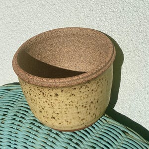 May include: A handmade ceramic bowl with a speckled yellow exterior and a cork rim. The bowl is sitting on a blue wicker surface, with a light blue wall in the background. The bowl is likely used for serving or storage.