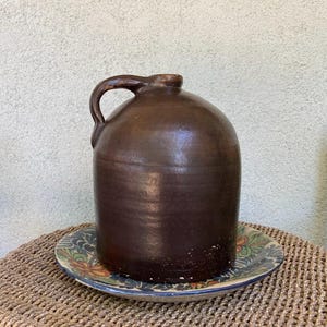 May include: A dark brown ceramic jug with a single side handle and a cylindrical body, resting on a round plate decorated with a colorful floral pattern. The jug features a smooth, glazed finish and a narrow neck with a small opening at the top.