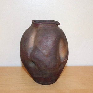 May include: A handcrafted, dark brown ceramic vase with a unique, organic shape. The vase has a wide, rounded body and a narrow opening. The surface has a textured appearance with subtle variations in color, creating a rustic aesthetic. The vase sits on a wooden surface.