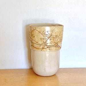 May include: A handmade ceramic vase with a two-tone design. The lower portion is a smooth, off-white color, while the upper section features a tan, textured surface with a delicate, botanical pattern. The vase has a cylindrical shape.