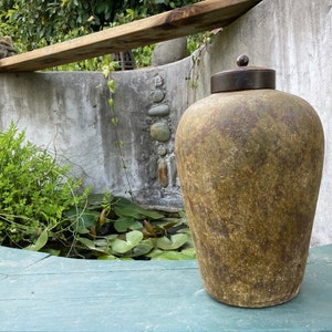 May include: A large, weathered, olive-green ceramic jar with a dark metal lid. The jar is cylindrical with a rounded top and sits on a green surface. The background includes a concrete wall, greenery, and a small pond.