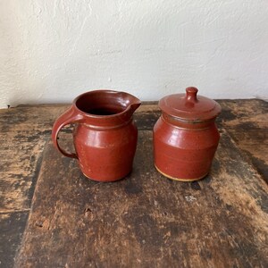 May include: A terracotta-colored ceramic creamer pitcher with a curved handle and a matching lidded sugar bowl. Both pieces feature a smooth, speckled finish with a slightly darker rim and a rounded, functional shape for serving beverages.