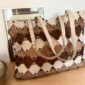 May include: A handcrafted tote bag with a unique geometric design. The bag features a patchwork of diamond-shaped pieces in shades of white, tan, and brown, secured with small, dark rivets. It has two handles and a rectangular shape.