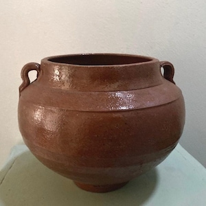May include: A brown ceramic pot with two curved handles. The pot has a rounded body and a wide opening. The exterior has a textured surface and a glossy finish. The pot is likely used for decorative purposes or storage.