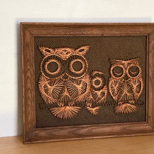 May include: A framed artwork featuring three owls crafted from copper-colored thread on a dark brown fabric background. The owls are arranged in a family grouping, with a wooden frame.