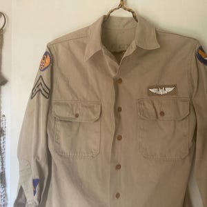 May include: Tan button-down shirt with two chest pockets and patches on the sleeves. The patches include a circular emblem, stripes, and a winged emblem. The shirt is hanging on a natural fiber hanger.