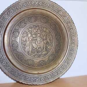 May include: A large, antique brass plate with intricate engraved designs. The plate features a central scene with figures and animals, surrounded by floral and geometric patterns. The rim has a scalloped edge.