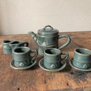 May include: A ceramic tea set in a muted blue-green glaze featuring a teapot with a lid, a creamer, and three cups with matching saucers. Each piece has a textured bamboo-inspired design with horizontal ridges and small triangular accents.