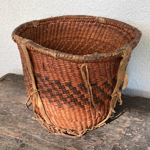 May include: A round, woven basket made of brown natural fibers featuring a dark brown geometric pattern around the center. The basket has a reinforced rim and vertical leather-like straps attached to the sides that extend to the base.