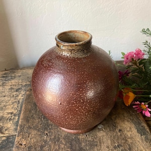 May include: A round, spherical ceramic vase with a dark brown, speckled glaze and a textured surface. The vase features a short, narrow neck with a slightly flared rim that has a lighter, mottled brown finish around the opening.