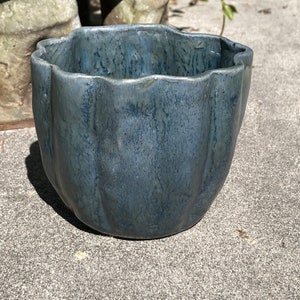 May include: A blue ceramic planter with a wavy rim. The planter has a textured surface and a dark blue glaze. The container is sitting on a concrete surface. The planter is suitable for indoor or outdoor use.