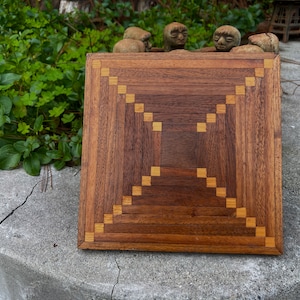 May include: A square wooden box lid featuring a geometric marquetry pattern. The design consists of dark brown wood slats arranged in a concentric square formation, accented by small, light-colored square inlays at the corners of each layer.